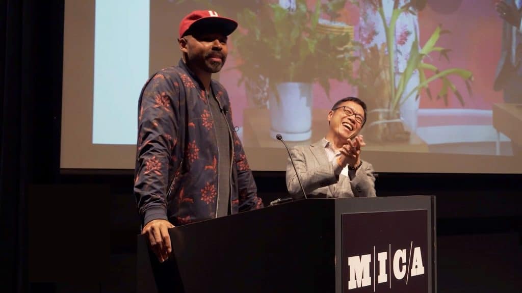 Hilton Carter and Sammy Hoi are standing at a podium with a sign on the front reading "MICA." Behind them is a screen with a presentation. Hilton is looking out to the audience, Sammy is clapping and smiling.