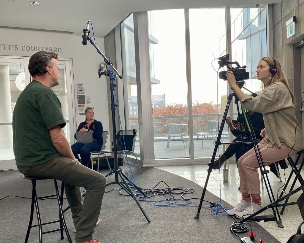 A spokesperson video filming session taken behind the scenes: we see a man in a green shirt seated in front of a woman behind a camera, who is filming him.