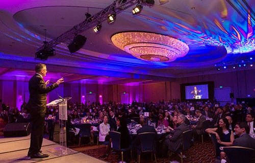 Photography Example: A well-dressed man stands on stage at a podium, addressing a large crowd seated at their tables at a gala.
