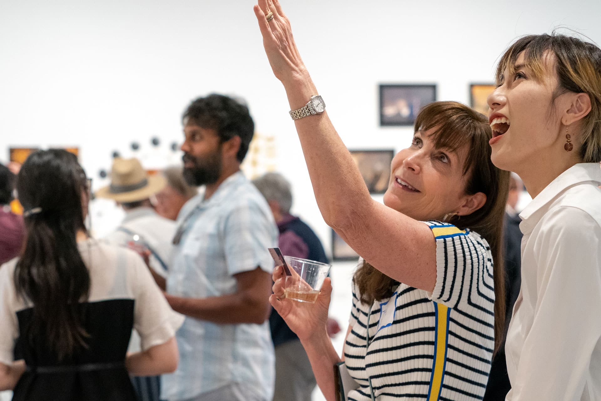 Highlight Video example: two women are in an art gallery. The woman in the background is wearing a striped shirt, and has long hair. She is pointing up at something. The woman in the forground has shorter hair, and she is smiling, also looking at what the other woman is pointing to.