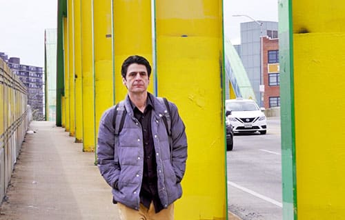 Video Production Example: a man dressed casually walks down the pedestrian sidewalk of a bridge, which is painted bright yellow. Cars are in the background. The man appears lost in thought, leaving the viewer to winder what he's thinking about, as an example of a micro documentary filming session.