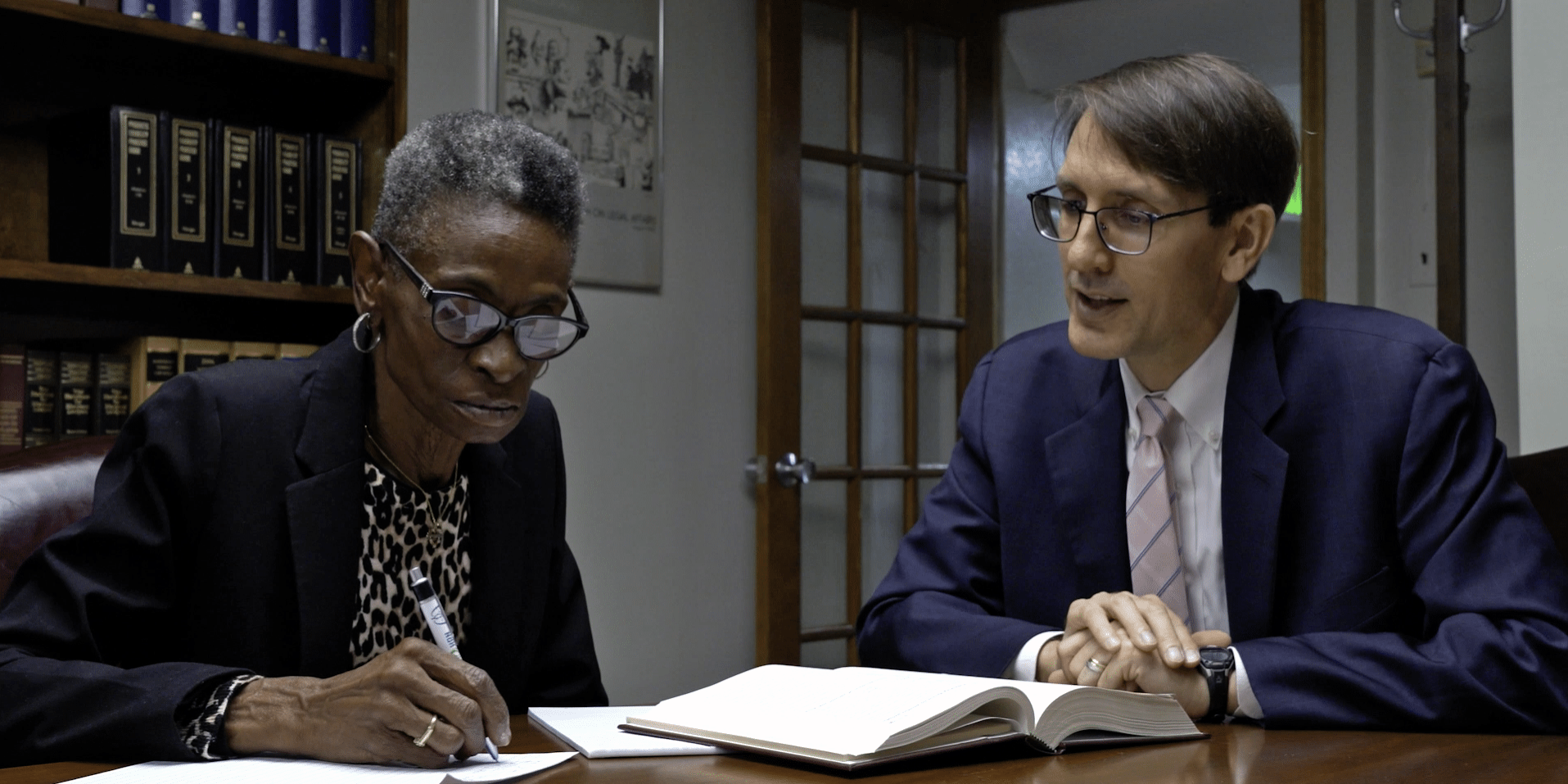 Micro Documentary Example: two individuals are seated at a table, an elderly black woman with gray in her hair, and a younger man with glasses. They are both dressed professionally. Papers and books are spread out on the table in front of them as they appear to be working together.